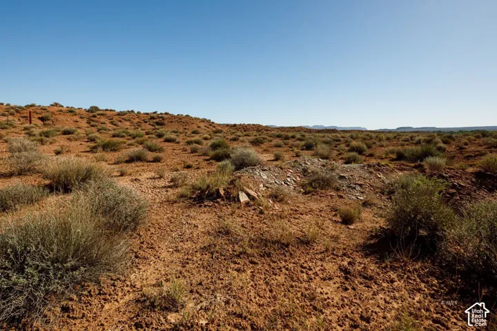 Expansive desert landscape featuring reddish-brown soil, sparse green vegetation, natural rock formations, and distant undulating terrain under a clear sky
