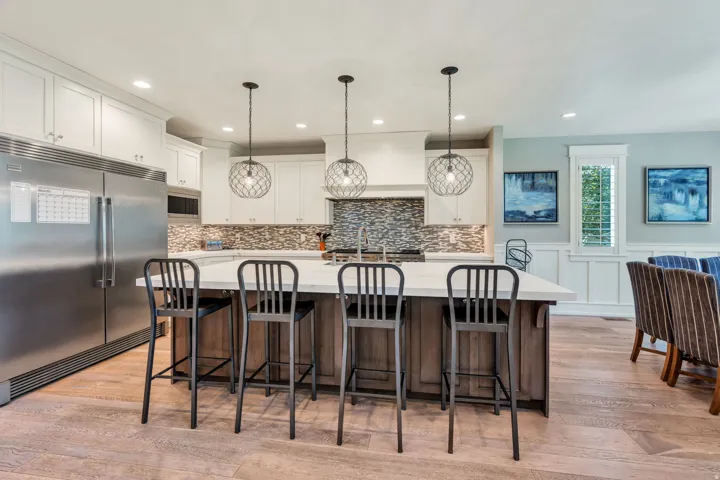 Kitchen featuring white upper cabinetry, a multi-tone tile backsplash, stainless steel appliances, and a central island with wood-finish base and light-toned countertop
