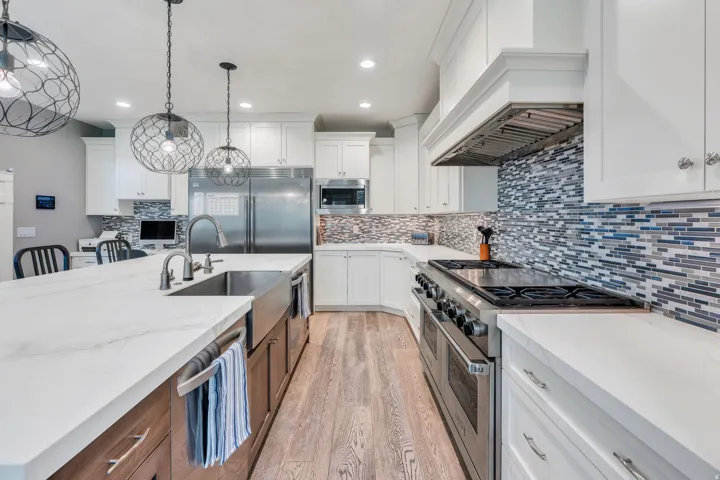 Spacious kitchen featuring an expansive island with a farmhouse sink, stainless steel appliances, white cabinetry, wood-finish flooring, and a linear tile backsplash