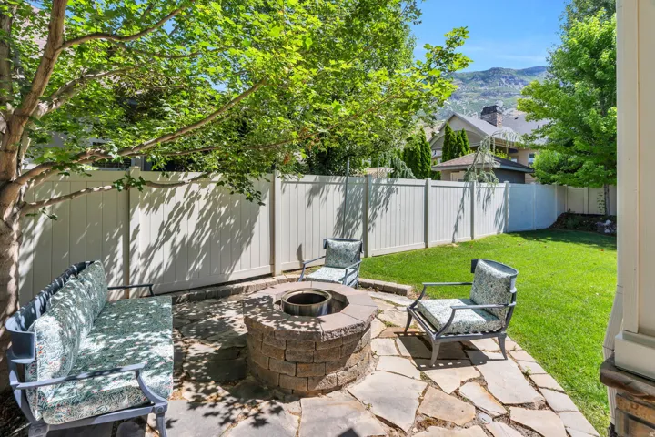Backyard patio featuring a stone-clad fire pit, irregular flagstone paving, a vibrant green lawn, mature trees, and a privacy fence