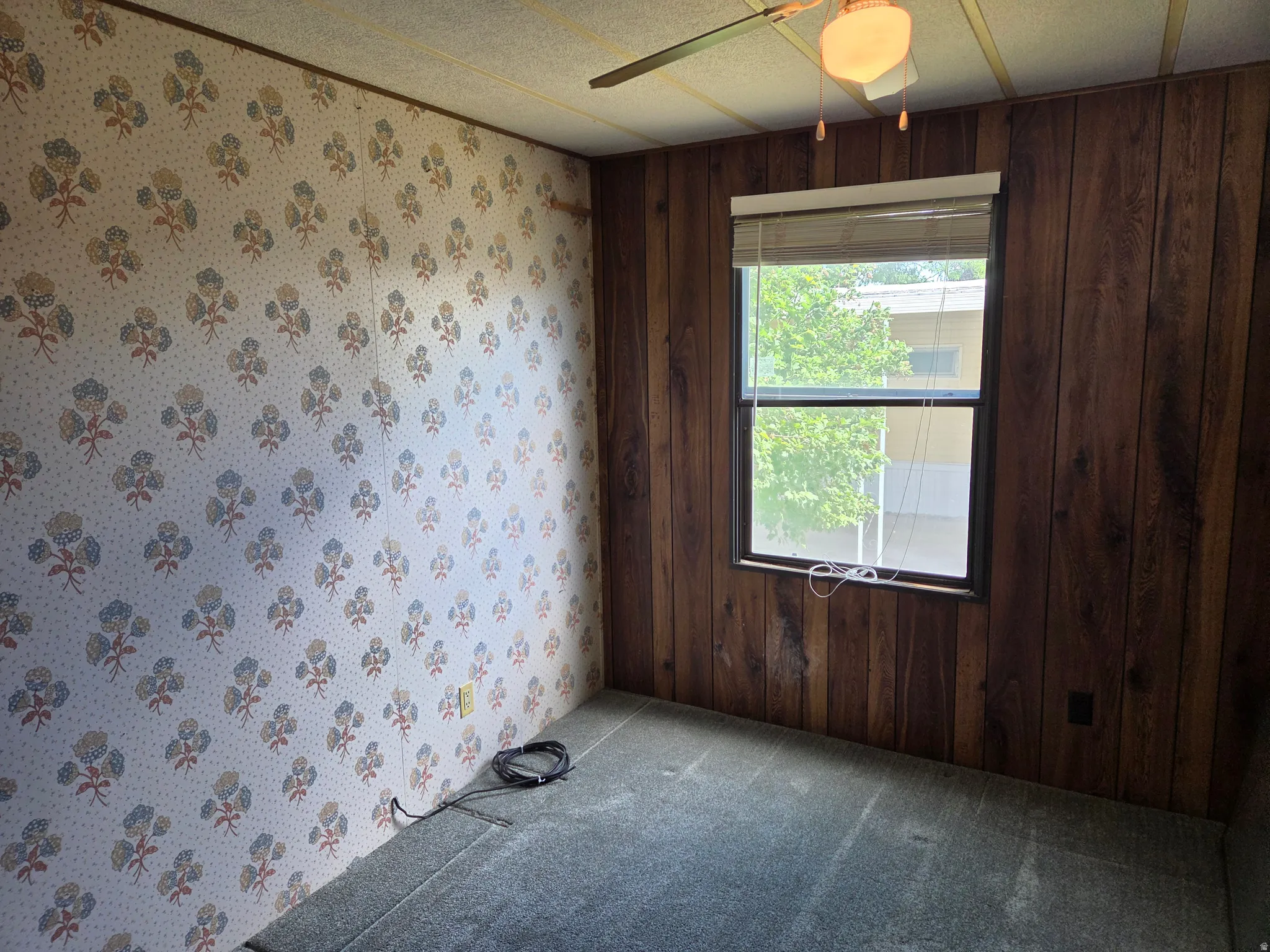 Room featuring a window with blinds, wood-paneled wall, patterned wallpaper, and ceiling fan with light fixture
