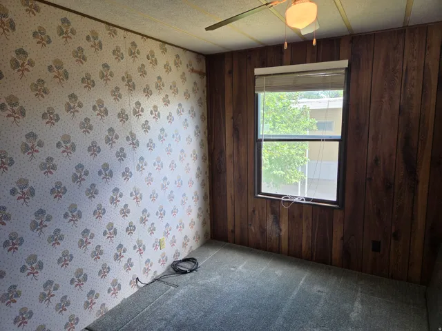 Room featuring a window with blinds, wood-paneled wall, patterned wallpaper, and ceiling fan with light fixture
