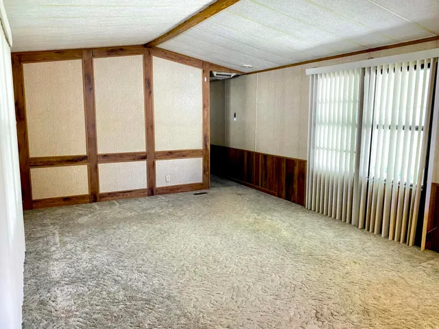 Spacious room featuring textured carpeting, a window with vertical blinds, and a paneled accent wall with wood-finish trim and wainscoting