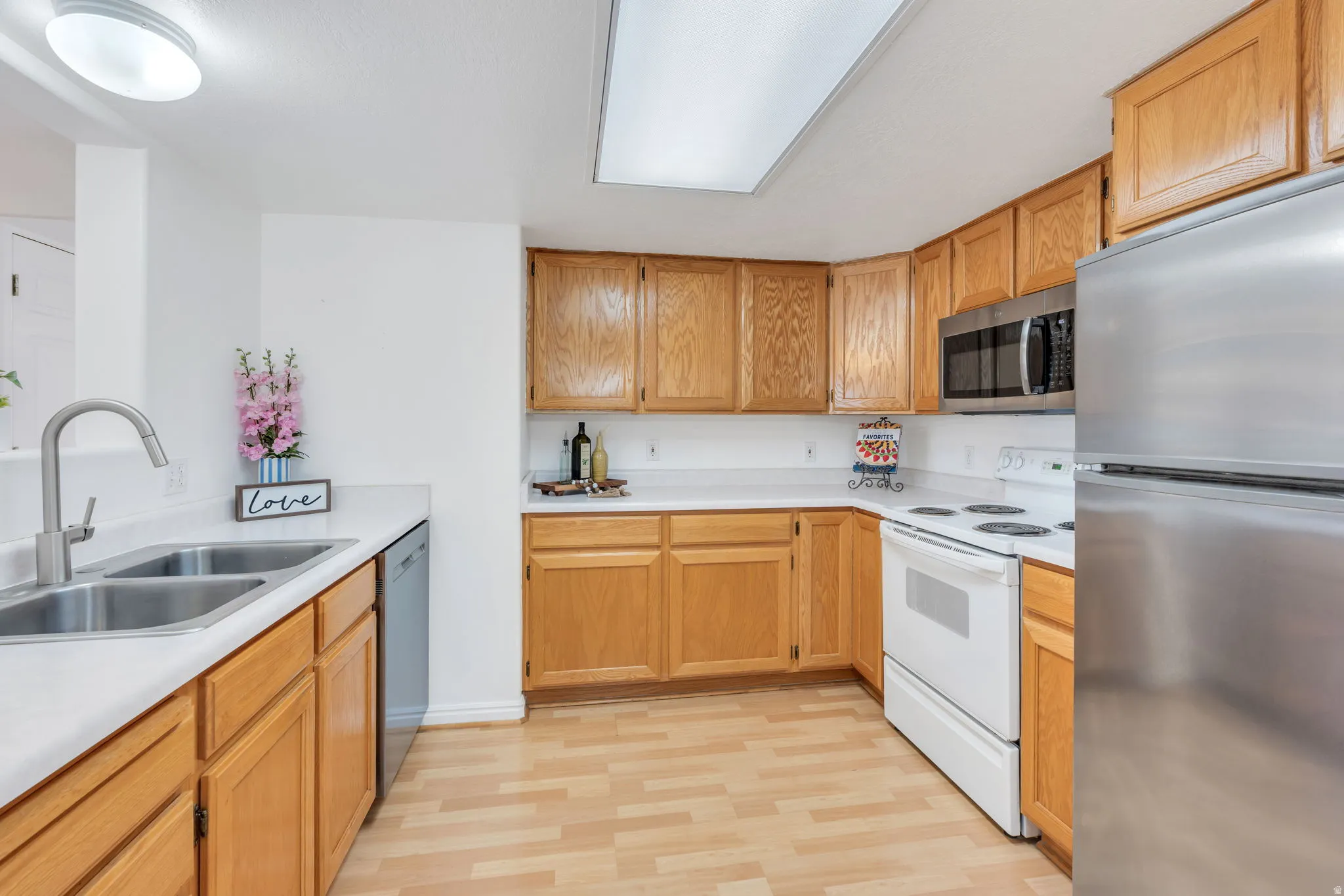 Open-concept kitchen featuring light-toned cabinetry, laminate countertops, a double basin sink with a gooseneck faucet, a stainless steel microwave, and wood-finish flooring