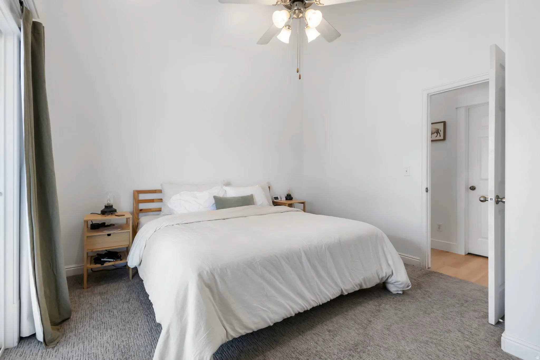 Carpeted room featuring a ceiling fan with light fixture, white walls, and a doorway leading to a space with wood-finish flooring