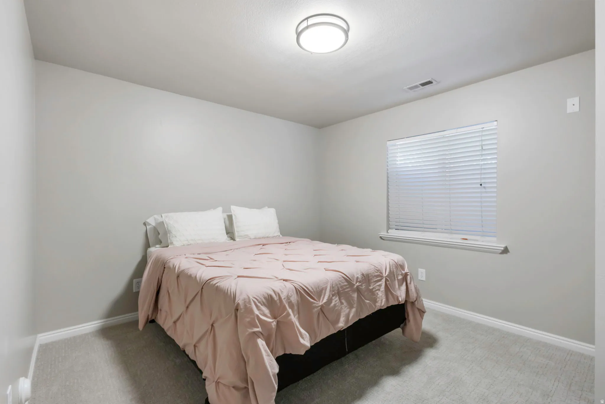 Comfortable room featuring light gray walls, plush carpeting, a single window with blinds, and a flush-mount ceiling light fixture