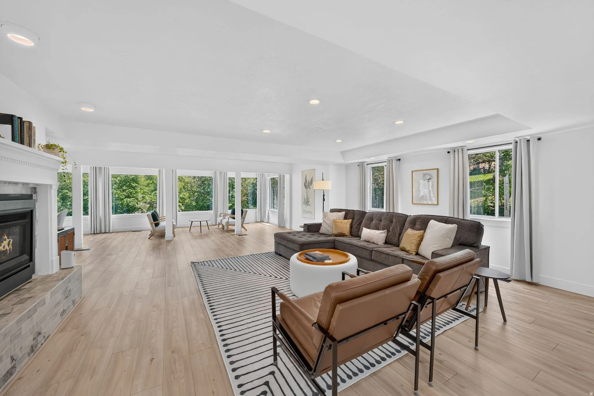 Spacious living area featuring light wood-finish flooring, a fireplace with a mantel and tiled hearth, recessed lighting, and expansive windows