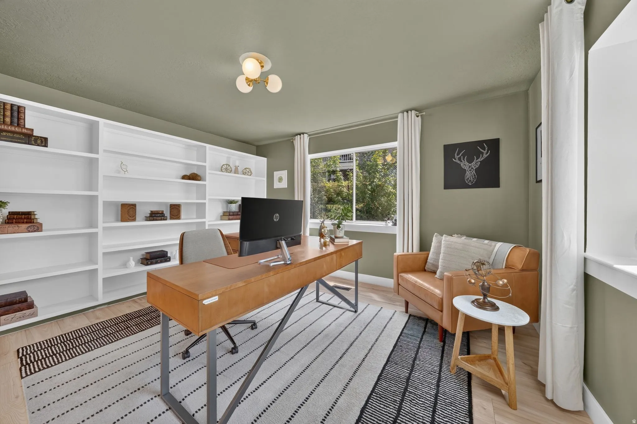 Dedicated office space featuring extensive built-in white shelving, wood-finish flooring, a window offering natural light, a modern ceiling light fixture, and neutral wall paint