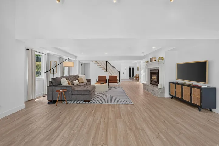 Spacious living area featuring wood-finish flooring, a stone-faced fireplace with a white mantel, and ample natural light from multiple windows