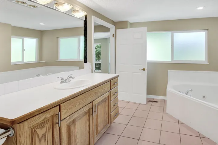 Ensuite Bathroom featuring a wood-finish vanity with a white countertop and integrated sink, a large wall-mounted mirror, a corner jetted tub, and frosted windows
