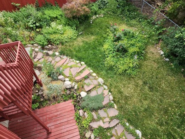 Expansive backyard featuring a red wood-stained deck with railing, a flagstone path, mature landscaping, and a manicured lawn area