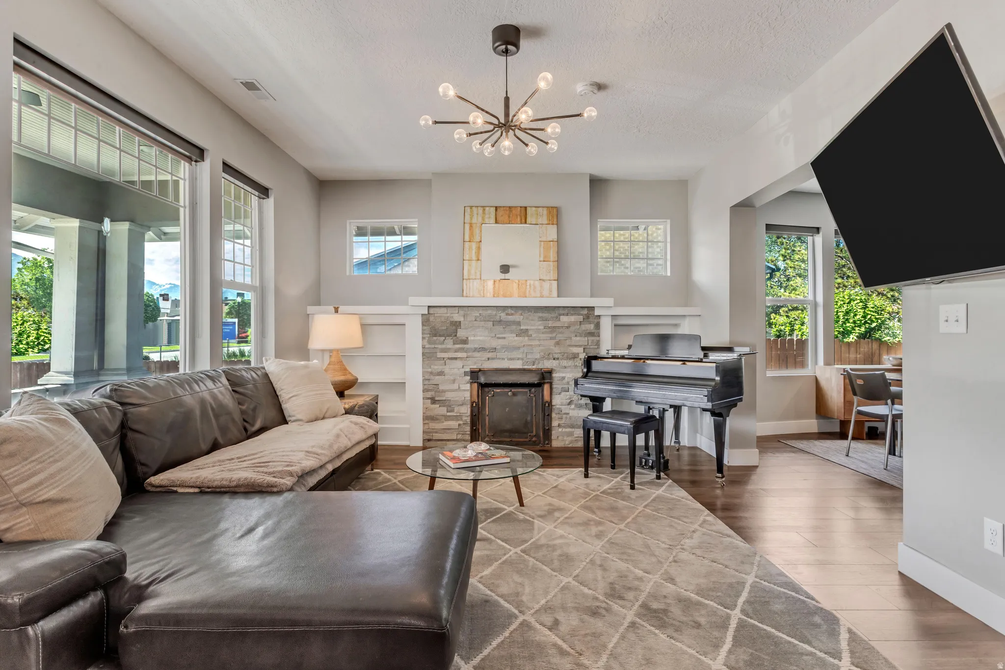 Spacious living area featuring wood-finish flooring, a stone-clad fireplace, built-in shelving, large windows with transom details, and a contemporary chandelier