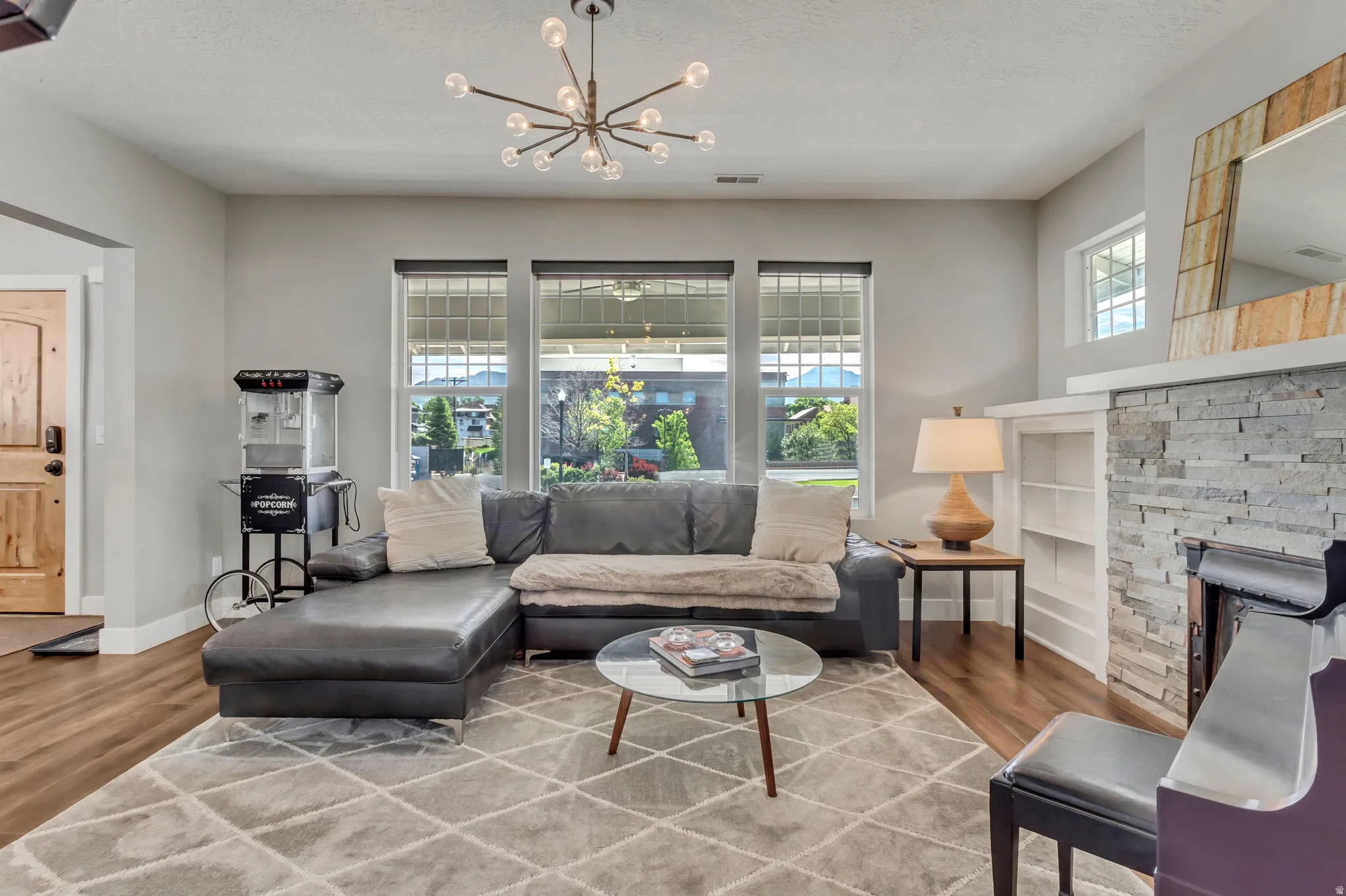 Living area featuring wood-finish flooring, a contemporary chandelier, a stone-clad fireplace with built-in shelving, and large windows with transom details