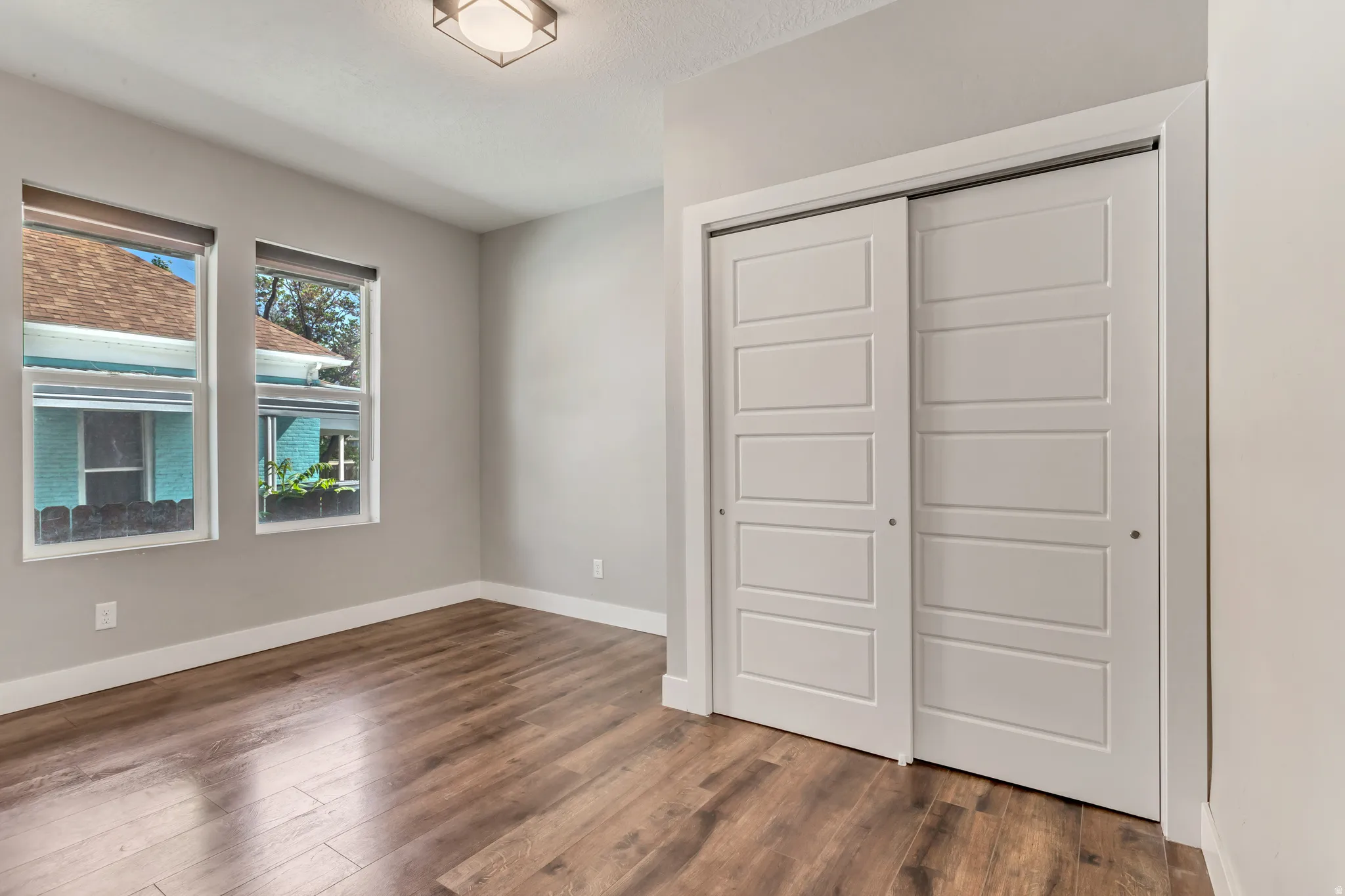 Spacious room featuring wood-finish flooring, two large windows, a flush-mount ceiling light fixture, and a sliding closet with paneled doors