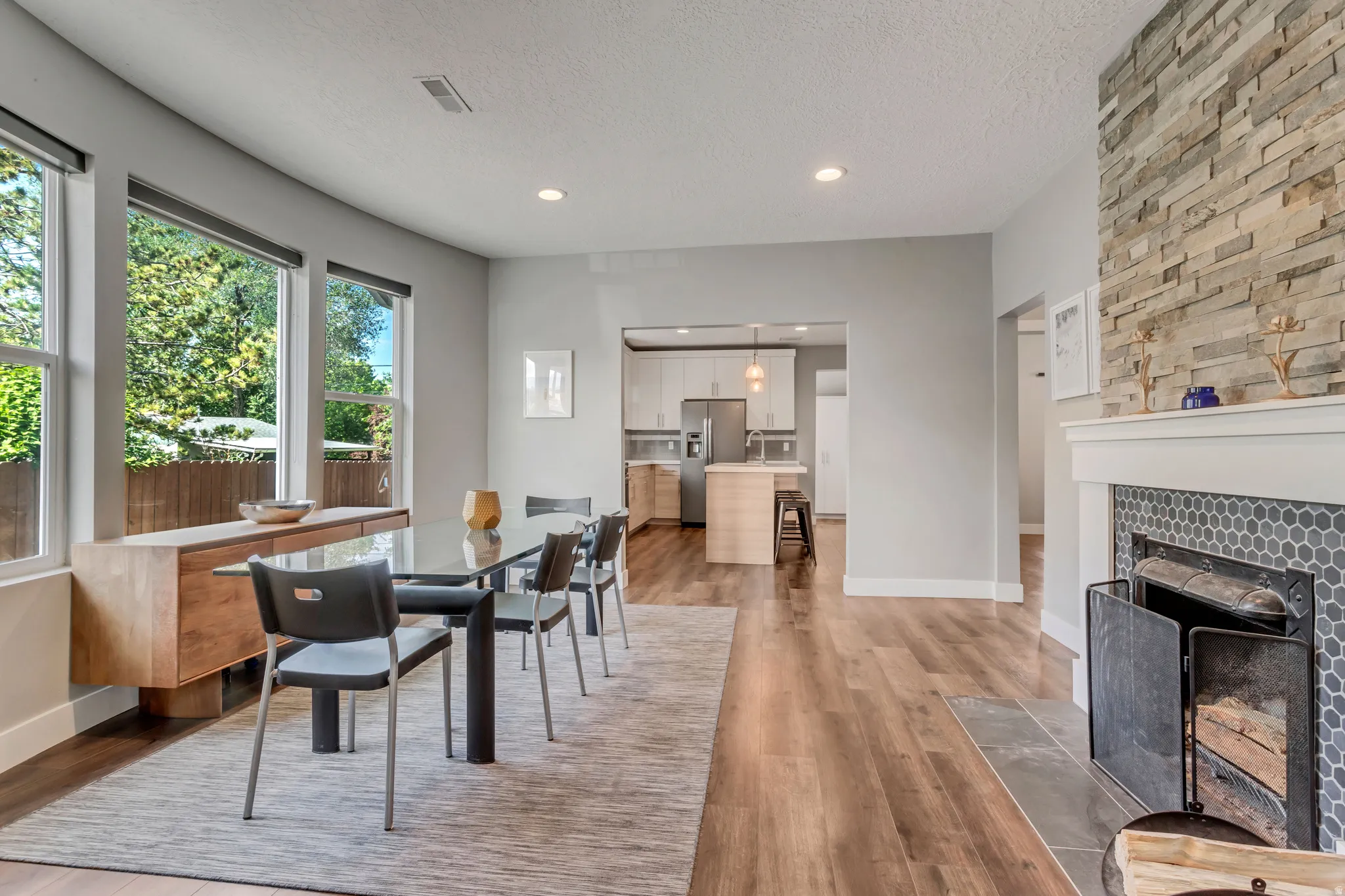 Spacious dining area featuring wood-finish flooring, a stacked stone fireplace with a hexagon tile surround and white mantel, and large windows providing natural light