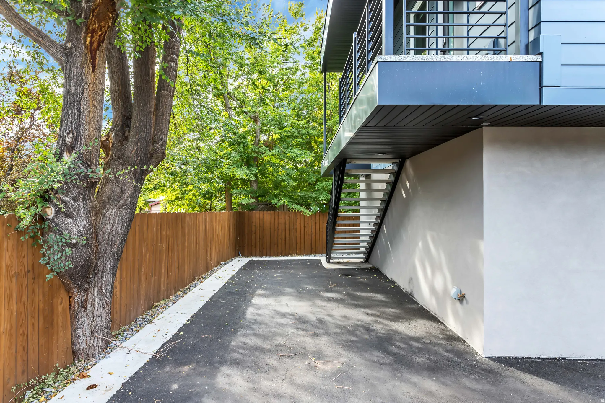 Paved driveway with a light-colored border, bordered by a wooden privacy fence and mature trees