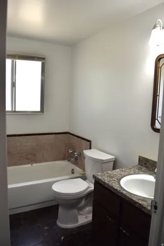 Full bathroom featuring a window, white bathtub with tile surround, white toilet, and a vanity with a white basin and stone-finish countertop