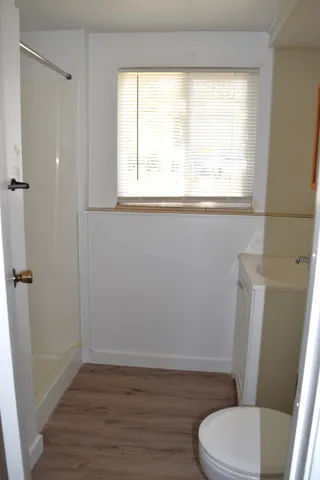Compact bathroom featuring a white single-piece shower, wood-finish flooring, white wainscoting, and a window with blinds