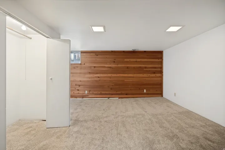 Spacious room featuring a wood-paneled accent wall, neutral carpeting, flush-mount lighting, and an integrated closet with bi-fold doors