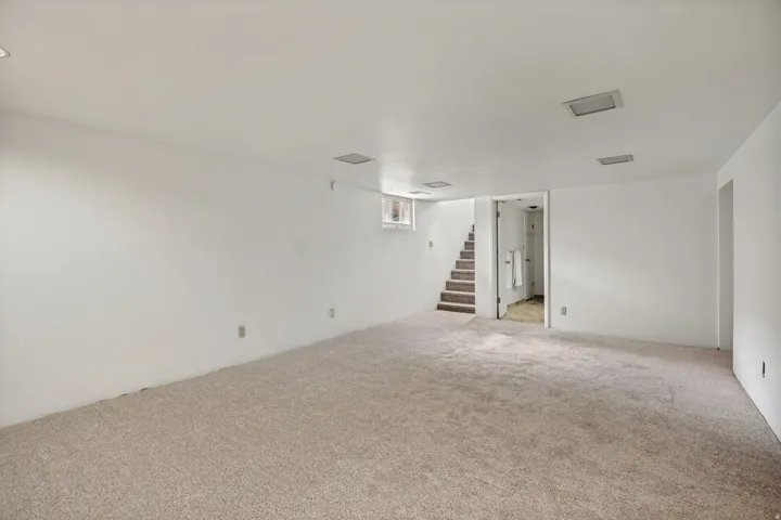 Expansive carpeted area with neutral-toned walls, recessed lighting, and a visible staircase providing access to an upper level