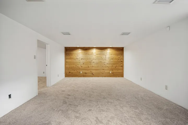 Spacious room featuring a prominent wood accent wall with recessed lighting, light-colored carpeting, white walls, and a doorway opening to an additional room