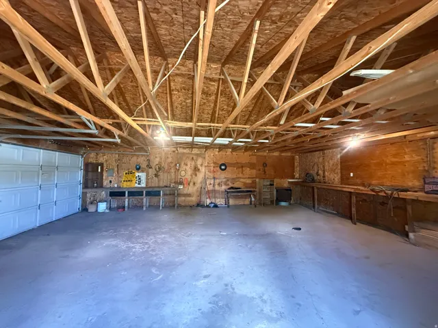 Spacious garage featuring an overhead door, exposed wood beam ceiling, concrete flooring, and extensive built-in wooden workbenches along the perimeter