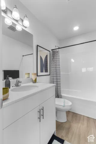 Contemporary bathroom featuring a white vanity with an integrated sink, a full-width mirror, a three-light vanity fixture, a white bath/shower combination, and wood-finish flooring