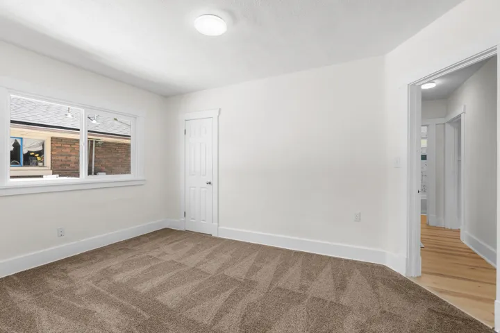 Spacious room featuring new carpet, crisp white walls, a large window with white trim, a solid door with white trim, and a ceiling-mounted light fixture