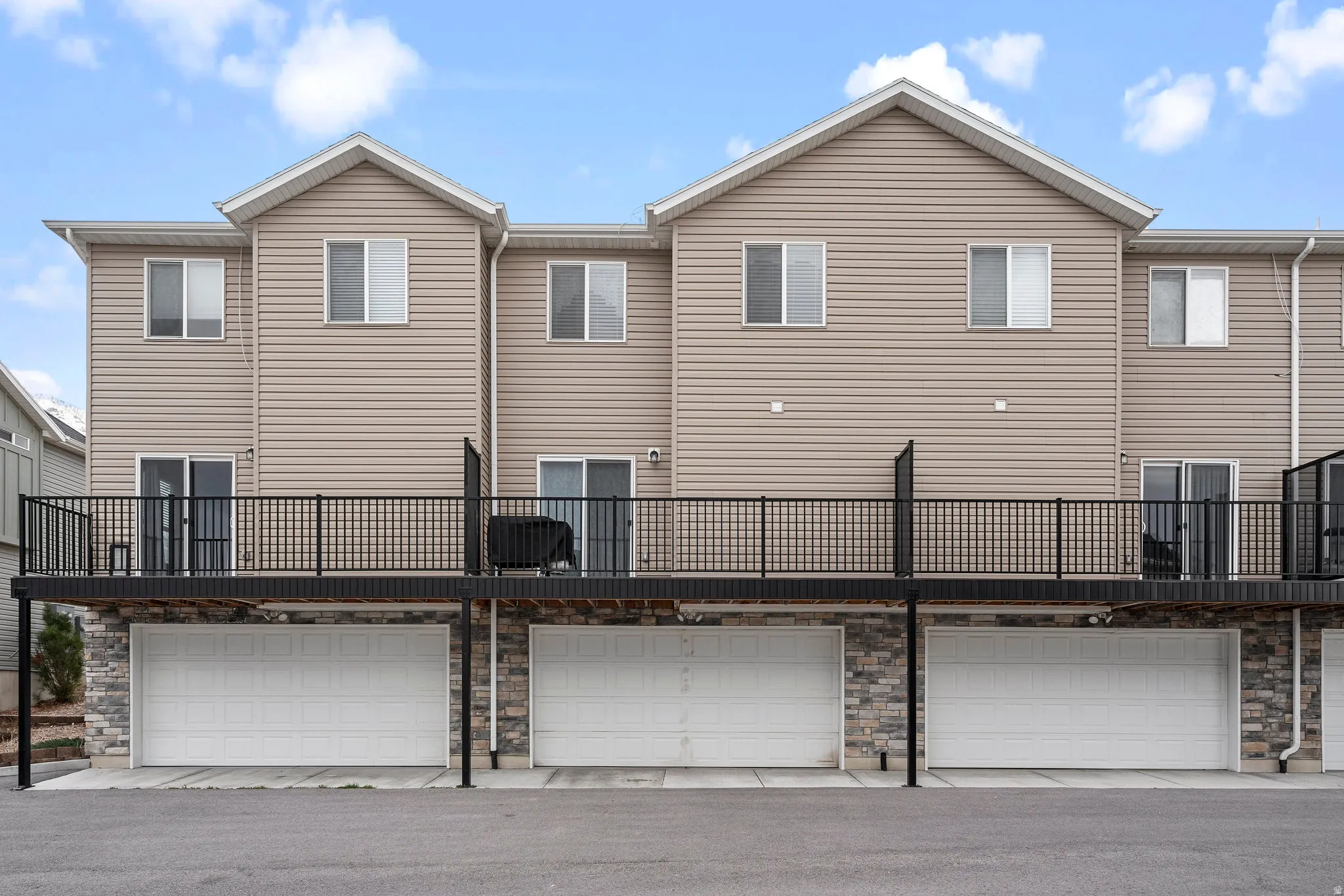 Multi-level exterior featuring tan siding, three individual garages with white doors, stone veneer accents, and a spacious deck with dark railing