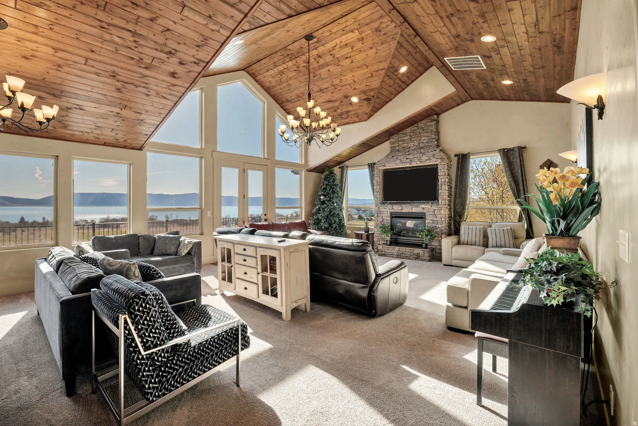 Spacious living area featuring a vaulted wood-paneled ceiling, stone-faced fireplace with hearth, recessed lighting, and expansive windows showcasing a prominent body of water and distant landforms