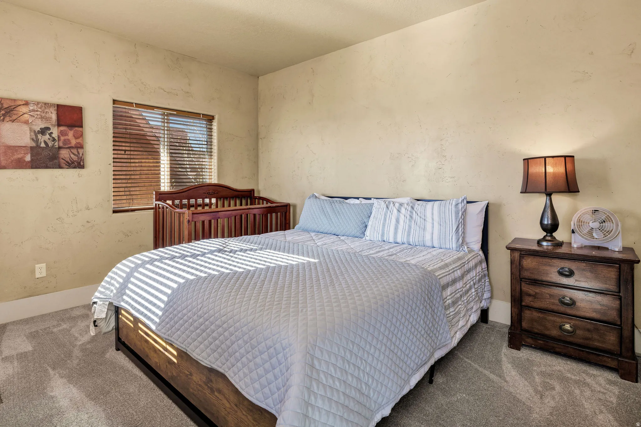 Bedroom featuring textured plaster walls, neutral-toned carpet flooring, and a window with wood-finish blinds