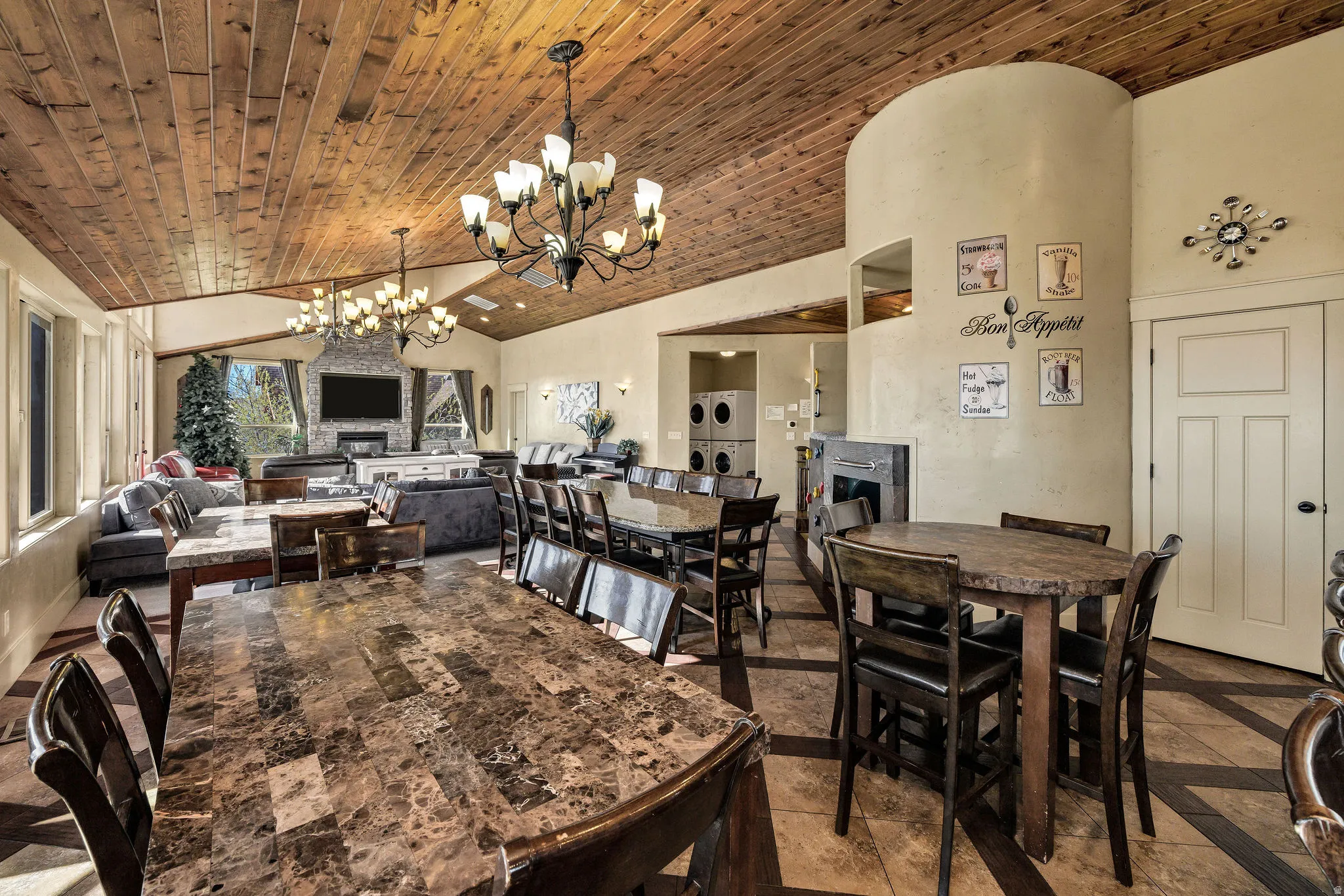 Vaulted wood-plank ceiling with multiple chandeliers, tile flooring, stone-front fireplace, large windows, and a built-in appliance stack