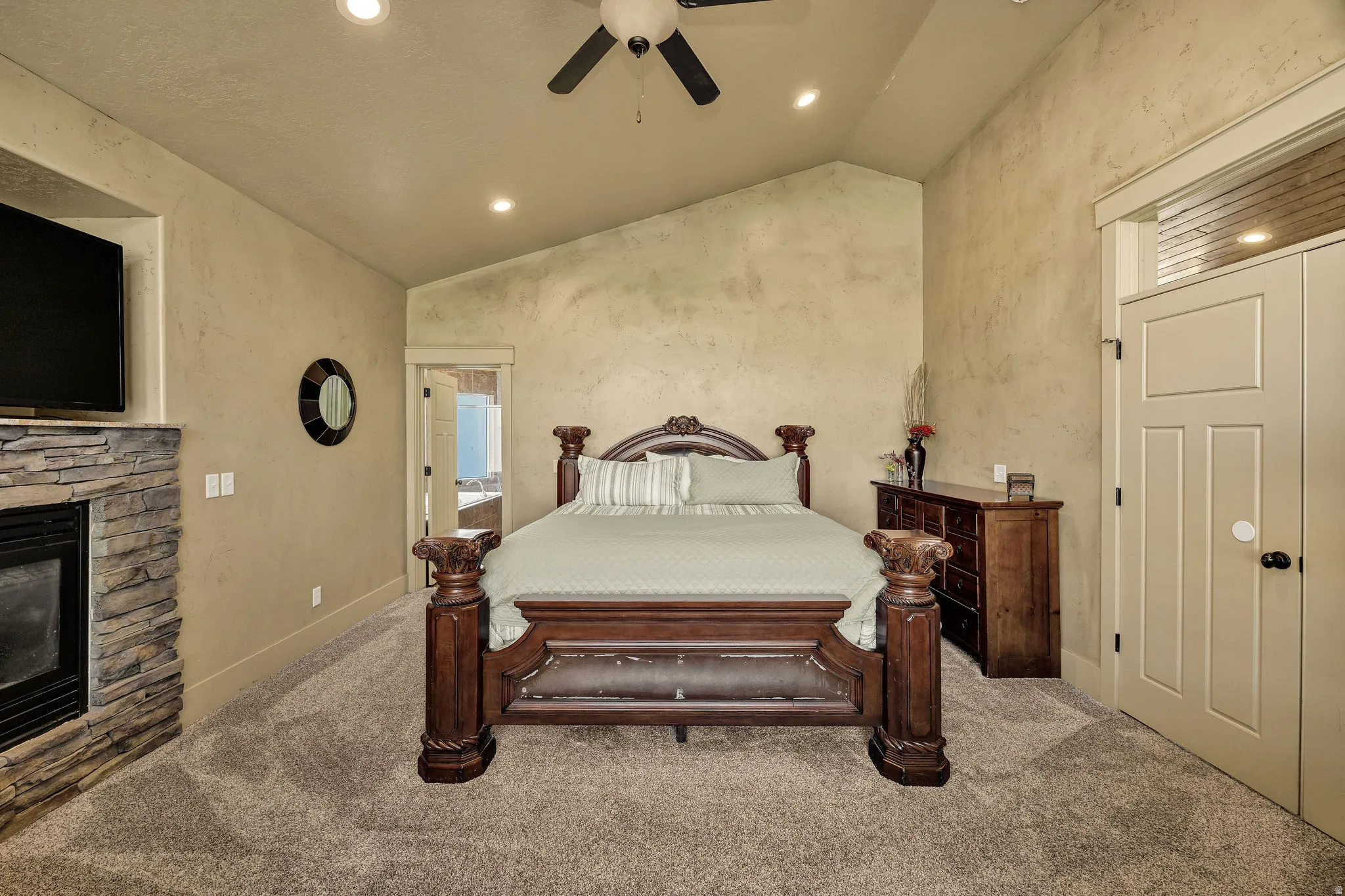 Vaulted ceiling bedroom featuring recessed lighting, a ceiling fan, plush carpet flooring, a stacked stone fireplace surround, and a paneled door with transom window