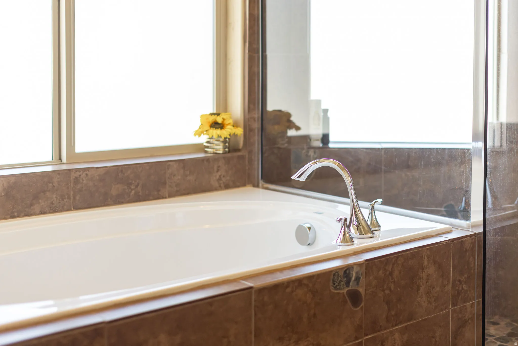 Soaking tub with polished chrome fixtures, surrounded by neutral-toned tile and a large window