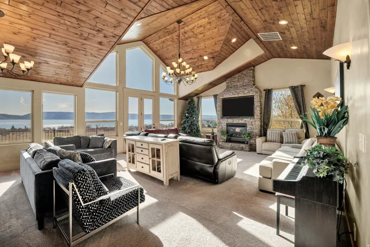 Spacious living area featuring a vaulted wood-paneled ceiling, stone-faced fireplace with hearth, recessed lighting, and expansive windows showcasing a prominent body of water and distant landforms