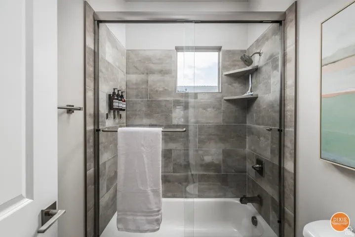Contemporary tub and shower combination featuring a glass enclosure with a sliding door, textured gray tile surround, built-in shelving, and a window providing natural light