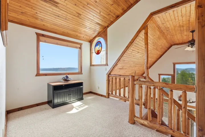 Carpeted loft area featuring a natural wood plank ceiling, log-style railings, and a large window with a scenic water backdrop