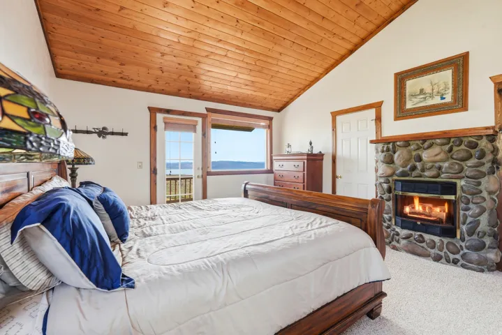Vaulted wood-paneled ceiling accentuates this room, featuring a stone-clad fireplace, a glass door leading to an exterior balcony, light carpet flooring, and wood trim detailing