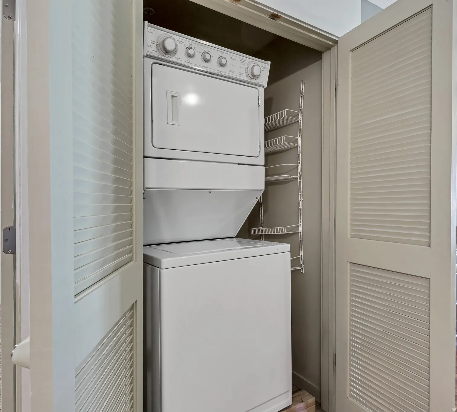 Dedicated laundry closet featuring a white stackable washer and dryer unit, louvered bi-fold doors, and integrated wire shelving