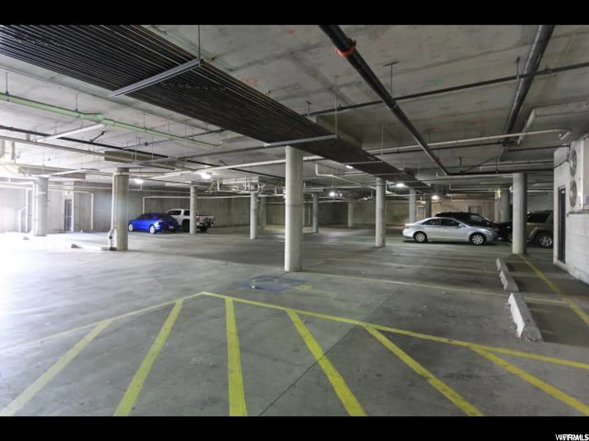 Concrete parking garage featuring ample marked spaces, exposed ceiling utilities, concrete support columns, and overhead lighting fixtures