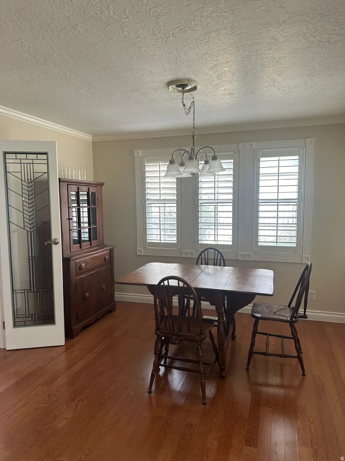 Wood-finish flooring, crown molding, a decorative chandelier, and three windows with plantation shutters