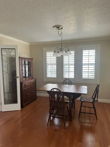 Wood-finish flooring, crown molding, a decorative chandelier, and three windows with plantation shutters