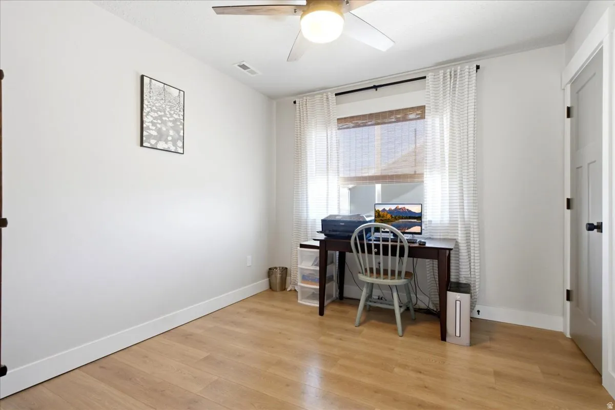 Bright room featuring wood-finish flooring, a window with natural light, a ceiling fan with integrated lighting, white walls, and a white interior door with black hardware