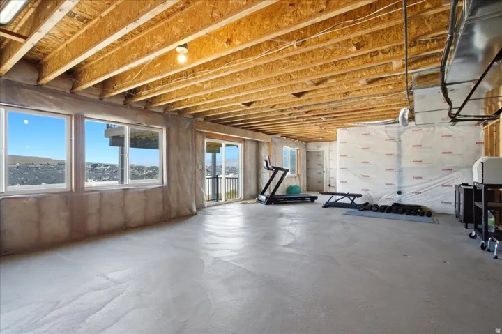 Daylight basement featuring three large windows, a sliding glass door with balcony access, exposed ceiling joists, concrete flooring, and an open layout