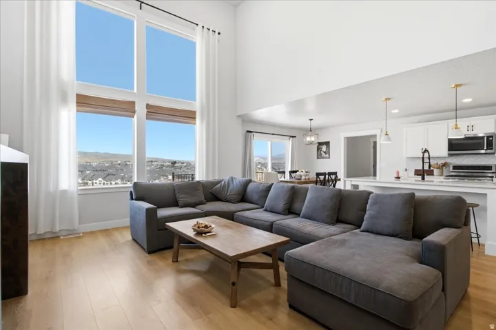 Great room featuring soaring ceilings, expansive window wall, light wood-finish flooring, and an open-concept kitchen with white cabinetry and pendant lighting