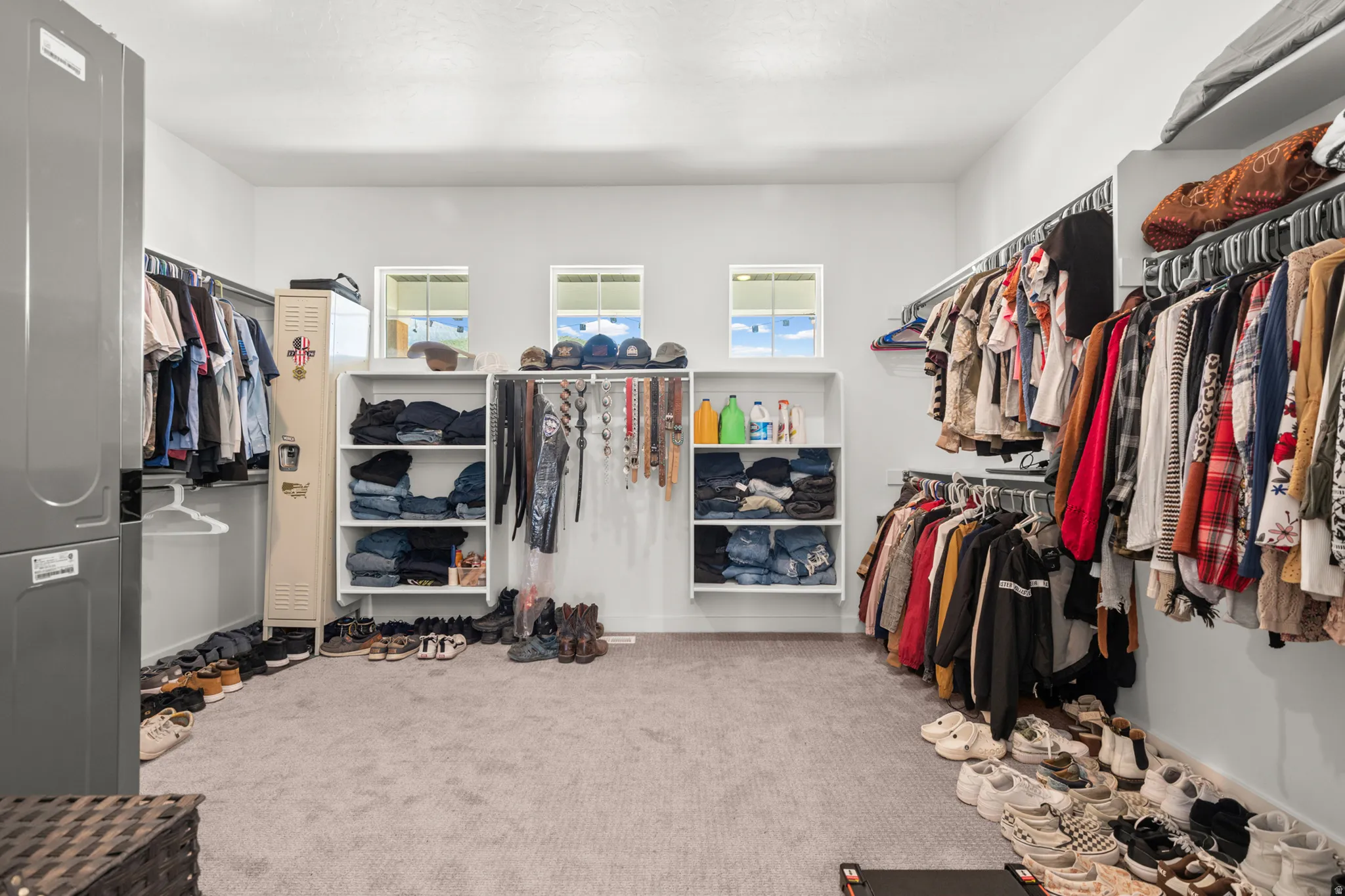 Spacious room featuring light gray carpeting, multiple built-in shelving units, extensive clothing racks, and three windows providing natural light