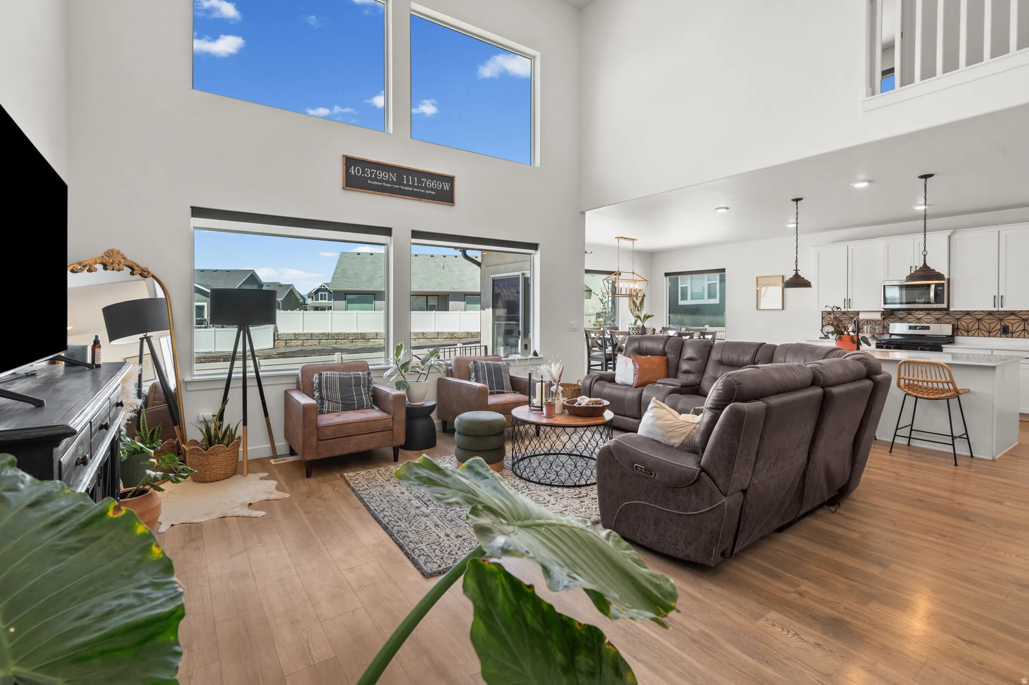 Spacious living area featuring high ceilings, wood-finish flooring, abundant natural light from large windows, an open-concept layout, and a modern kitchen with white cabinetry and stainless steel appliances