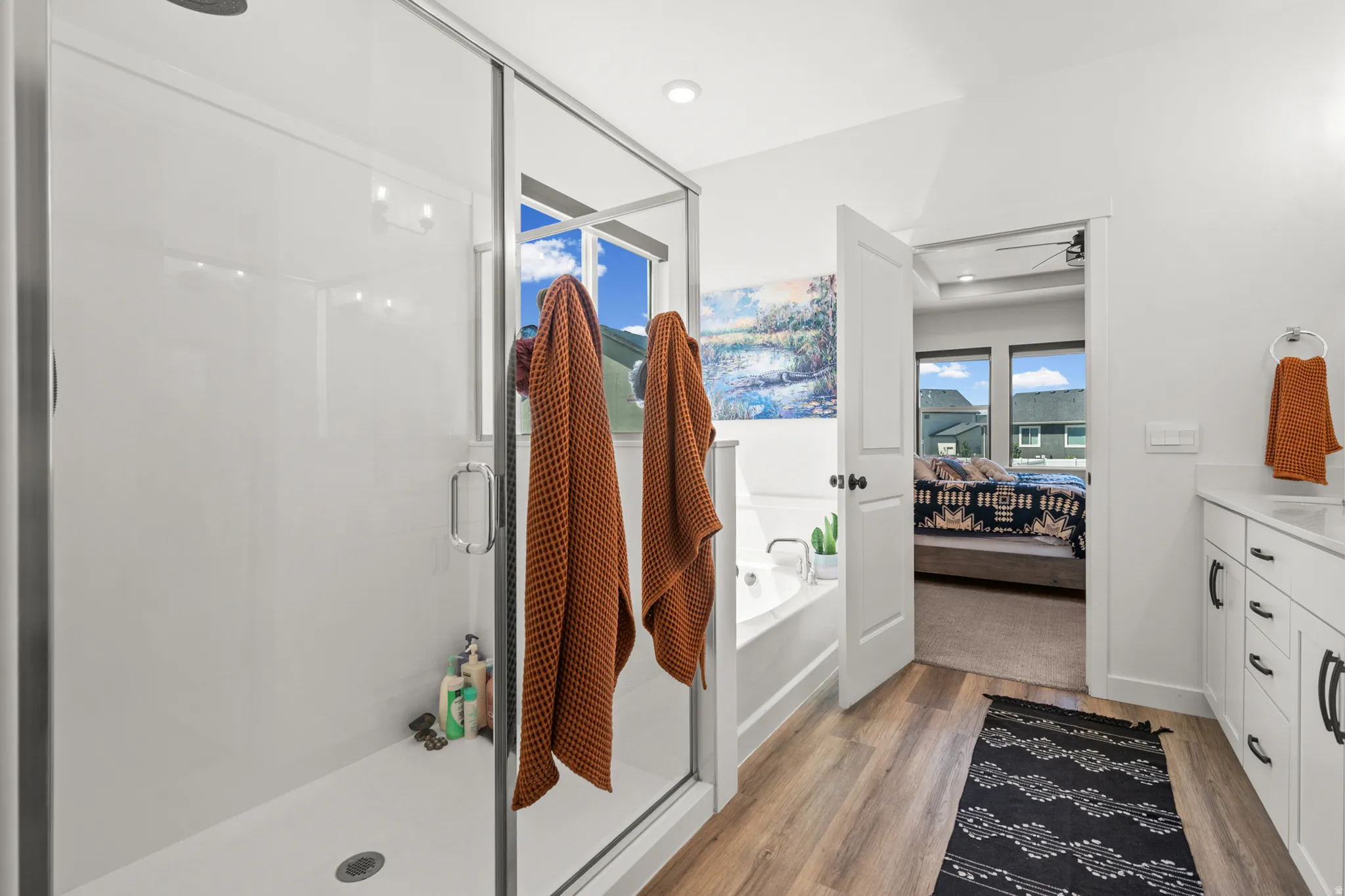 Glass-enclosed shower with a built-in bench, a soaking tub, wood-finish flooring, a white vanity with a white countertop, and dark-finish hardware
