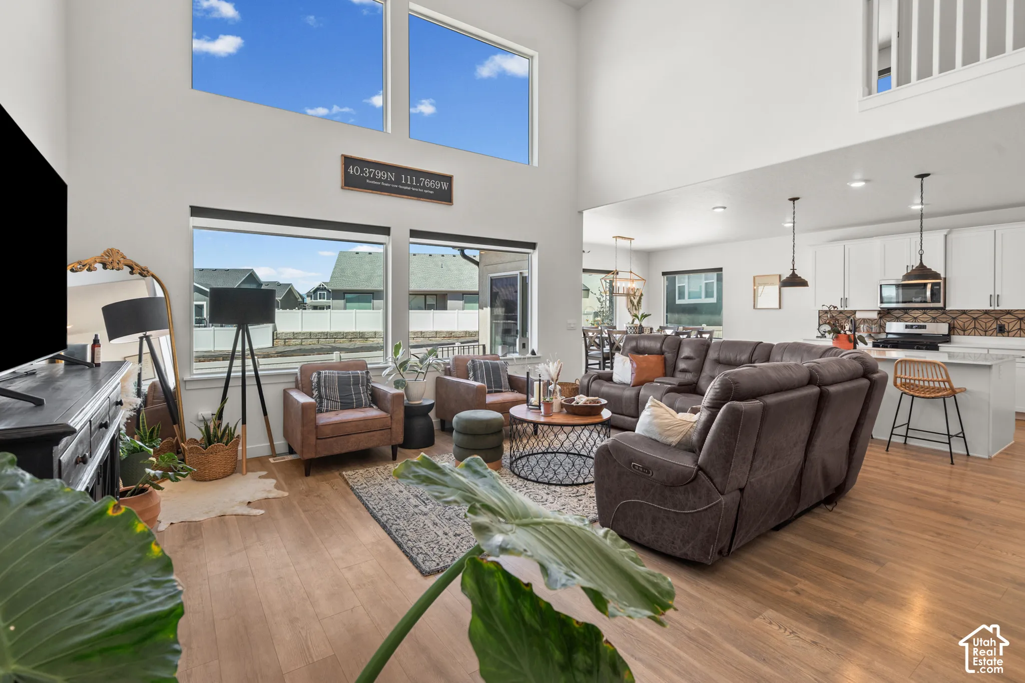Spacious living area featuring high ceilings, wood-finish flooring, abundant natural light from large windows, an open-concept layout, and a modern kitchen with white cabinetry and stainless steel appliances
