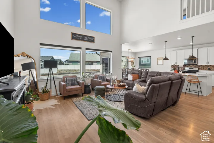 Spacious living area featuring high ceilings, wood-finish flooring, abundant natural light from large windows, an open-concept layout, and a modern kitchen with white cabinetry and stainless steel appliances